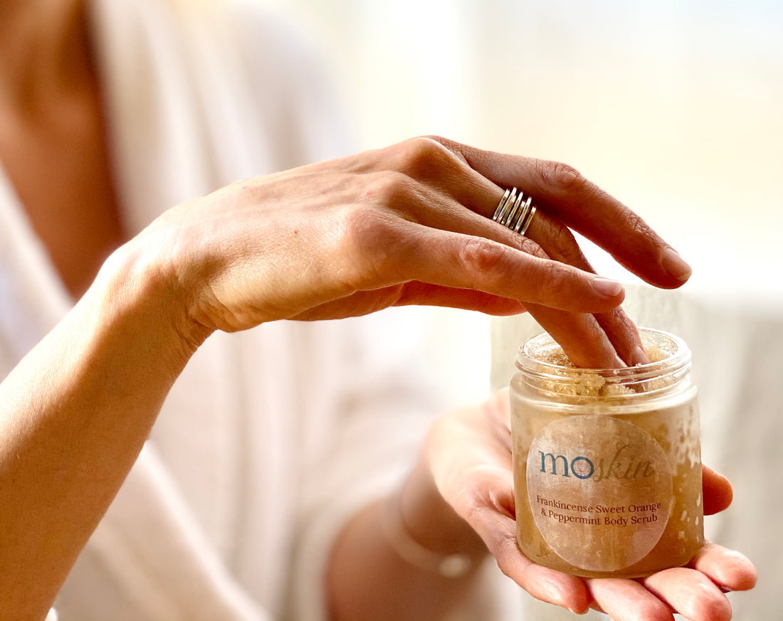 Hands massaging a sugar body scrub into a forearm in the shower, with water beading on skin and a soft towel nearby.