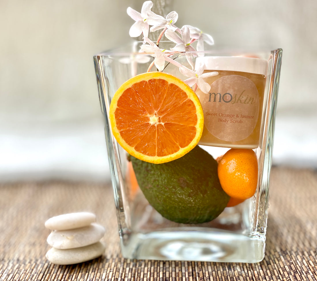 A morning bathroom scene with a citrus body scrub beside a jar of lemongrass body butter, fresh orange slices, and lemongrass stalks on a clean towel.