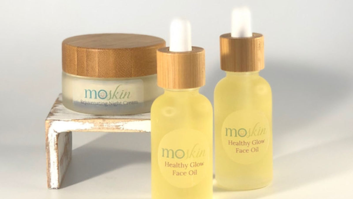 A clean, softly lit scene of a gua sha stone beside a bottle of Healthy Glow Oil with a few drops on the back of a hand, showing the ideal light slip for facial massage.