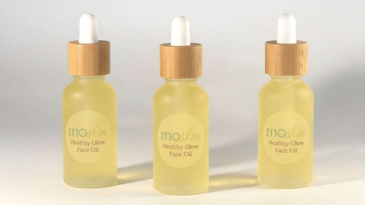 A close‑up of an oily or acne‑prone cheek with two small drops of a lightweight facial oil being pressed into damp skin with fingertips.