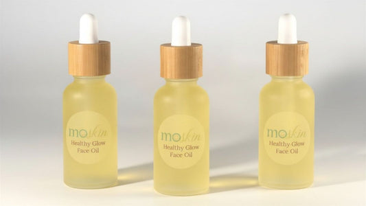 A close‑up of an oily or acne‑prone cheek with two small drops of a lightweight facial oil being pressed into damp skin with fingertips.