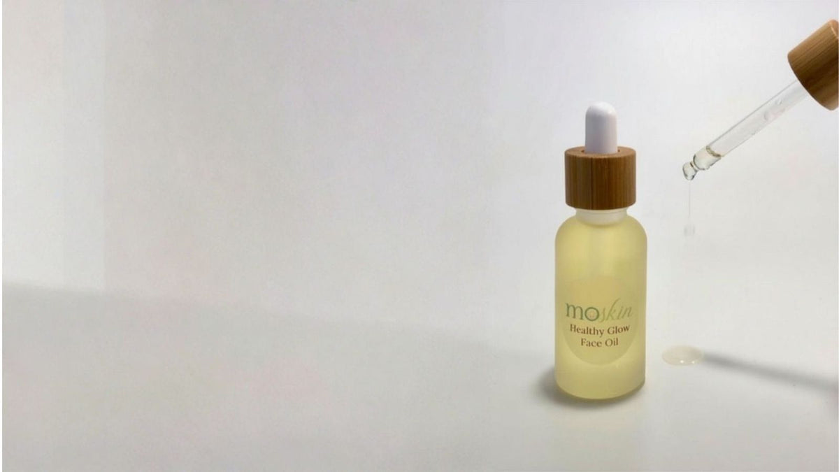 A few dewy drops of face oil being pressed into freshly cleansed, towel-damp skin beside a tube of sunscreen in morning light.