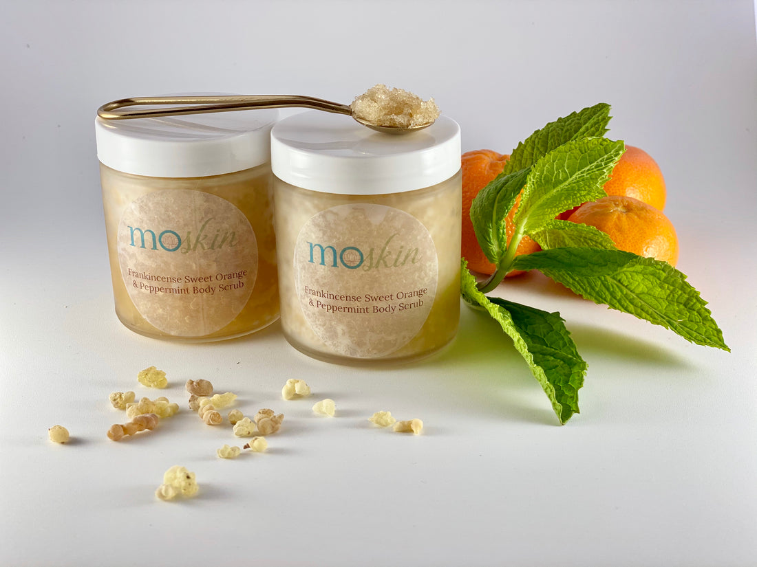 Close-up of a hand gently exfoliating a dry elbow, with a small wooden spoon of sugar scrub and a jar of body butter nearby.